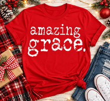 Load image into Gallery viewer, A red short-sleeve graphic T-shirt with the phrase 'amazing grace.' in distressed white lettering, styled in a holiday flat lay with jeans, white sneakers, a plaid blanket, pinecones, and Christmas decorations.