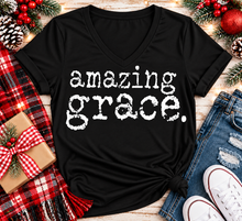 Load image into Gallery viewer, A black V-neck t-shirt with the words 'amazing grace.' printed in a distressed white typewriter font, styled in a flat lay with jeans, white sneakers, a red plaid scarf, a wrapped gift box, and Christmas pinecones and berries.