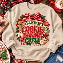 Load image into Gallery viewer, A beige crewneck sweatshirt laid flat on a rustic wooden surface, featuring a large festive graphic that reads 'Christmas Cookie Baking Crew' in red and green lettering with gingerbread cookies, a red bow, pine branches, and baking tools. The sweatshirt is styled with holiday props including a red plaid blanket, wrapped gift boxes, a plate of decorated cookies, and a red mug of hot cocoa.