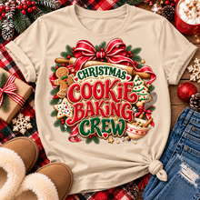 Load image into Gallery viewer, A beige graphic t-shirt with a festive Christmas cookie baking crew design, styled in a flat lay with holiday decorations, slippers, jeans, and a mug of popcorn.