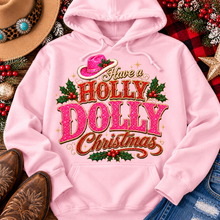 Load image into Gallery viewer, Have a Holly Dolly Christmas western graphic shirt featuring pink and red glitter-inspired lettering, a festive pink cowgirl hat, gold accents, and Christmas holly berries. Available in beige and pink in short sleeve crewneck, V-neck, long sleeve crewneck, and hoodie styles.