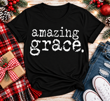 Load image into Gallery viewer, A black short-sleeve graphic T-shirt laid flat on a rustic wood surface, printed with the phrase 'amazing grace.' in distressed white lettering.