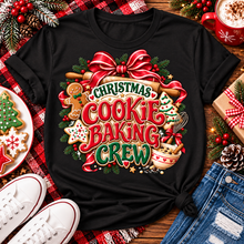 Load image into Gallery viewer, A black short-sleeve graphic T-shirt laid flat on a rustic wooden surface with holiday props. The shirt features a large festive cookie-baking crew design with red ribbon, green pine branches, gingerbread cookies, a Christmas tree, and the text 'Christmas Cookie Baking Crew.

