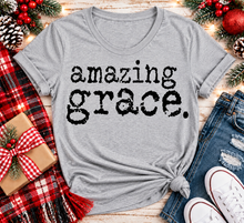 Load image into Gallery viewer, A heather gray short-sleeve t-shirt with 'amazing grace.' printed in black distressed text across the front, styled in a flat lay with jeans, white sneakers, a red plaid scarf, pinecones, and holiday decorations.
