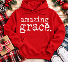 Load image into Gallery viewer, A red pullover hoodie with the words 'amazing grace.' printed in white distressed lettering across the chest, styled in a holiday flat-lay with a plaid scarf, gift box, pinecones, ornaments, jeans, and white sneakers on a wooden surface.
