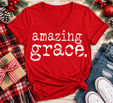 Load image into Gallery viewer, A red V-neck t-shirt with the words 'amazing grace.' printed in a distressed white font, styled in a flat lay with jeans, white sneakers, a plaid scarf, a wrapped gift, pinecones, and holiday berries on a light wood background.
