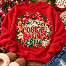 Load image into Gallery viewer, A red crewneck sweatshirt laid flat on a rustic wooden surface, featuring a large festive graphic that reads 'Christmas Cookie Baking Crew' with holiday illustrations including a gingerbread man, cookie, tree, ornaments, and baking tools. The sweatshirt is styled with holiday greenery, lights, boots, denim, and dessert props around it.
