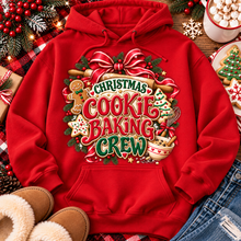 Load image into Gallery viewer, A red pullover hoodie with a large festive Christmas cookie baking crew graphic, styled in a cozy holiday flat-lay with jeans, slippers, gingerbread cookies, and holiday decorations.

