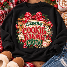 Load image into Gallery viewer, A black crewneck sweatshirt laid flat on a rustic wooden surface, featuring a large festive graphic that reads 'Christmas Cookie Baking Crew' in red, green, and gold lettering, surrounded by holiday illustrations like a gingerbread man, cookie, ribbon, and mixing bowl. The sweatshirt is styled with red plaid fabric, brown slippers, denim, and a mug of hot chocolate.

