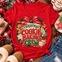 Load image into Gallery viewer, A red short-sleeve V-neck T-shirt laid flat on a rustic holiday surface, featuring a large Christmas cookie baking graphic with the words “CHRISTMAS COOKIE BAKING CREW,” cookies, gingerbread, ornaments, pinecones, and a red bow. Surrounding props include jeans, slippers, a mug of hot cocoa with marshmallows, wrapped gift boxes, and cookies.
