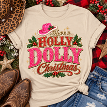 Load image into Gallery viewer, Have a Holly Dolly Christmas western graphic shirt featuring pink and red glitter-inspired lettering, a festive pink cowgirl hat, gold accents, and Christmas holly berries. Available in beige and pink in short sleeve crewneck, V-neck, long sleeve crewneck, and hoodie styles.
